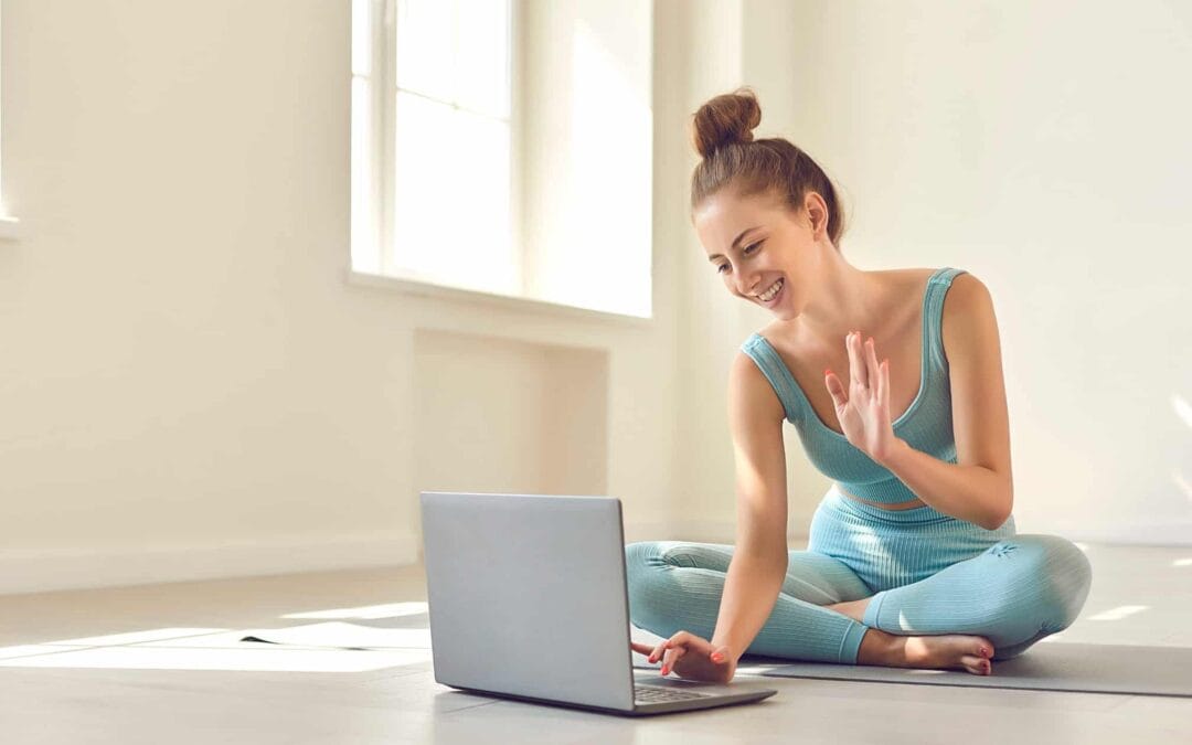 Femme entrepreneure assise en tailleur en tenue de sport, souriante, faisant un appel vidéo sur son ordinateur portable depuis chez elle. Elle symbolise une coach bien-être ou holistique, en interaction digitale. Image illustrant la création d’un site web aligné à son activité de coaching en toute simplicité.
