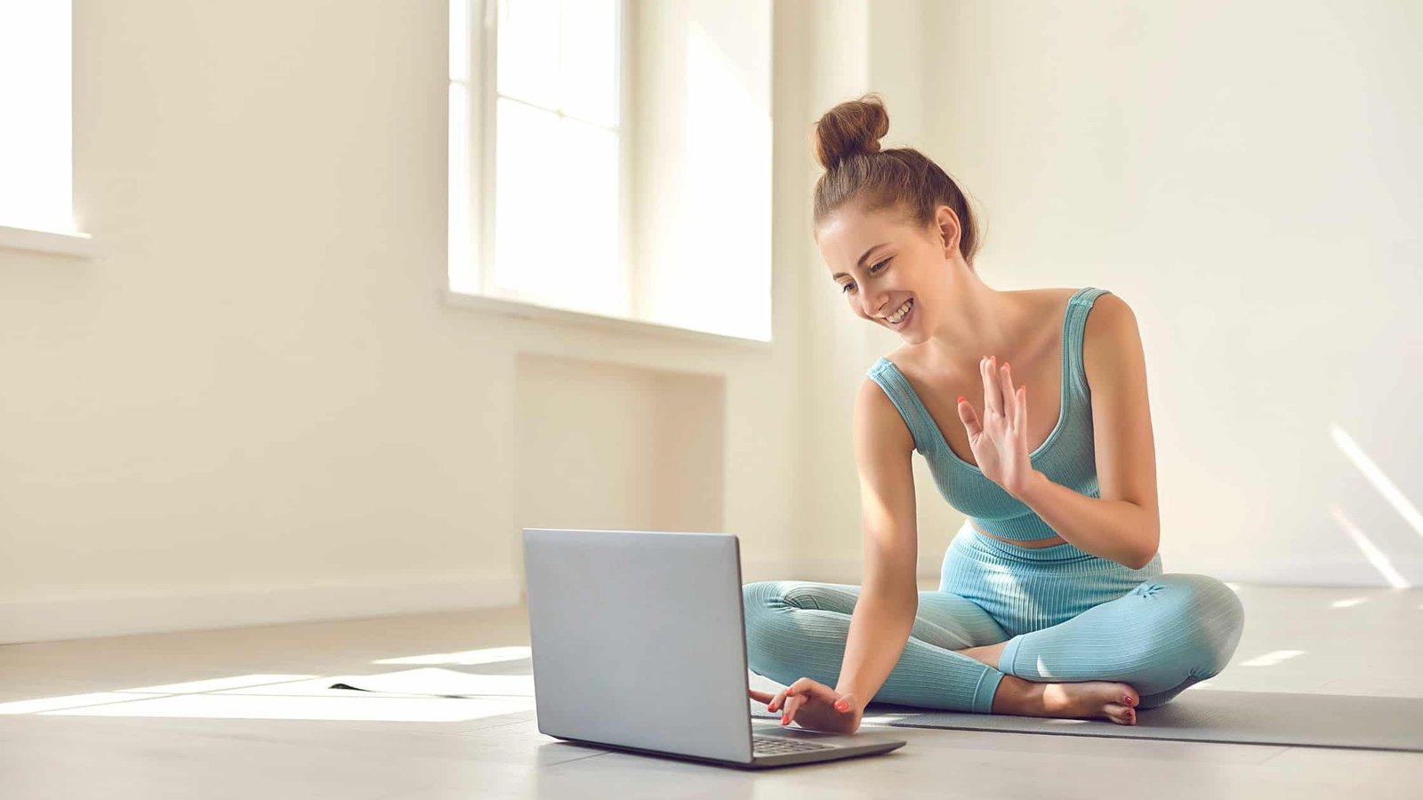 Femme entrepreneure assise en tailleur en tenue de sport, souriante, faisant un appel vidéo sur son ordinateur portable depuis chez elle. Elle symbolise une coach bien-être ou holistique, en interaction digitale. Image illustrant la création d’un site web aligné à son activité de coaching en toute simplicité.